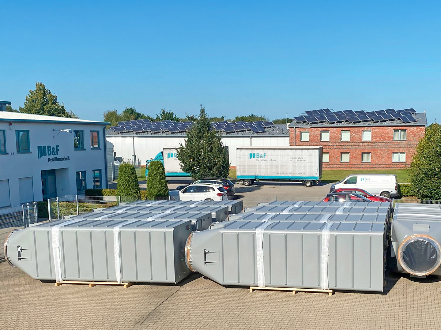 Mehrere große, graue Anlagenbehälter auf einem Betriebshof, im Hintergrund Firmengebäude und Fahrzeuge.