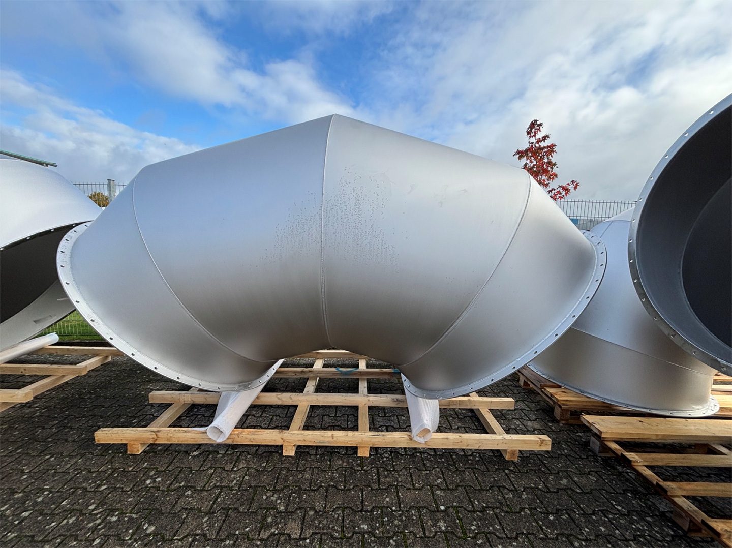 Großformatiges metallisches Rohrleitungsbogen-Bauteil auf Holzgestell vor blauem Himmel.