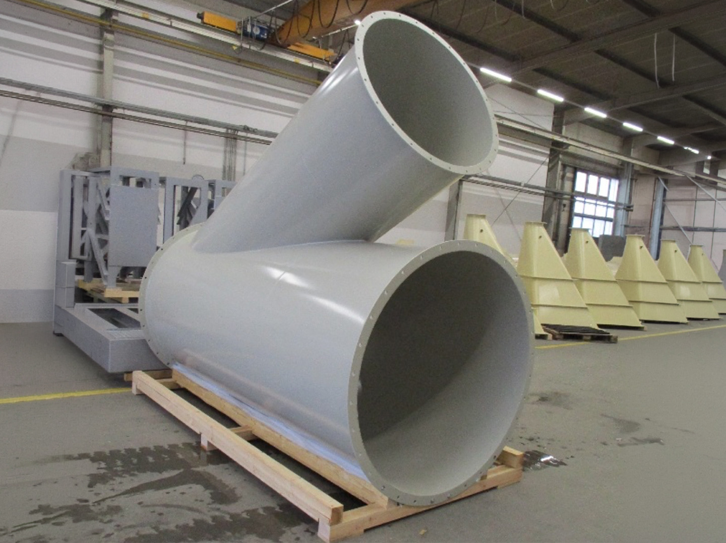 Großes metallisches Rohrleitungs‑Abzweigstück mit zwei schrägen Auslässen, gelagert auf einer Holzpalette in einer Werkhalle.